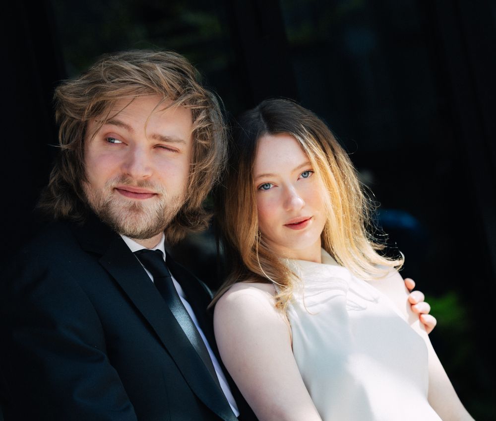 A young couple poses intimately outdoors, the man in a black suit and the woman in a white dress.