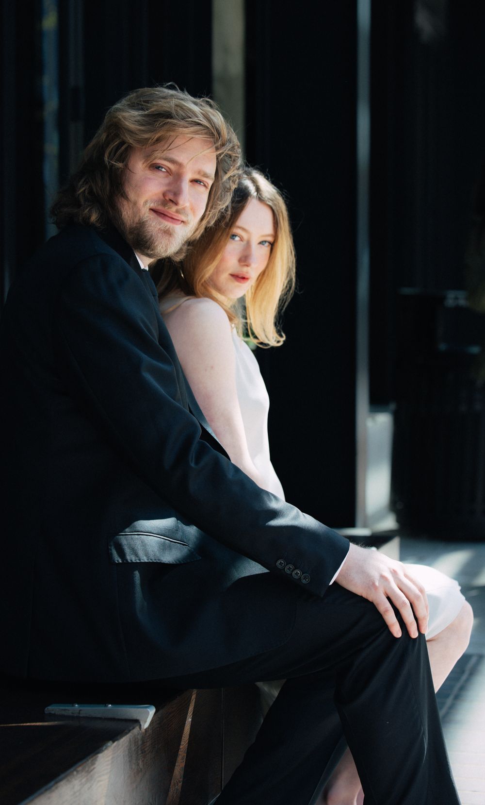 A couple sits closely together in sunlight, the man in a black suit and woman in a white dress.