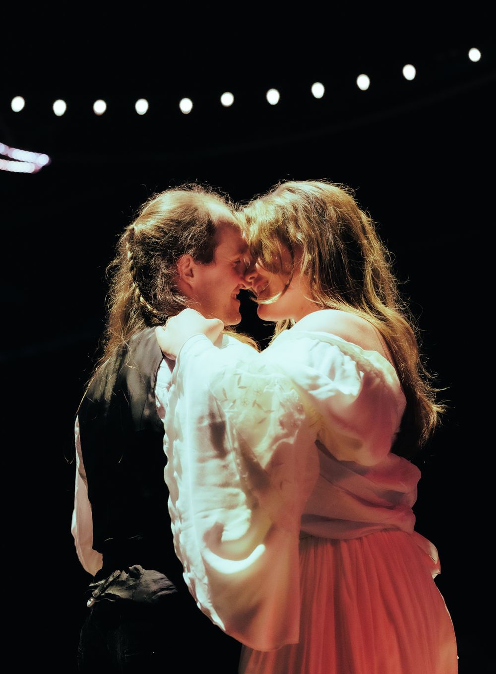 Two performers sharing a romantic moment on a theatrical stage under string lights.