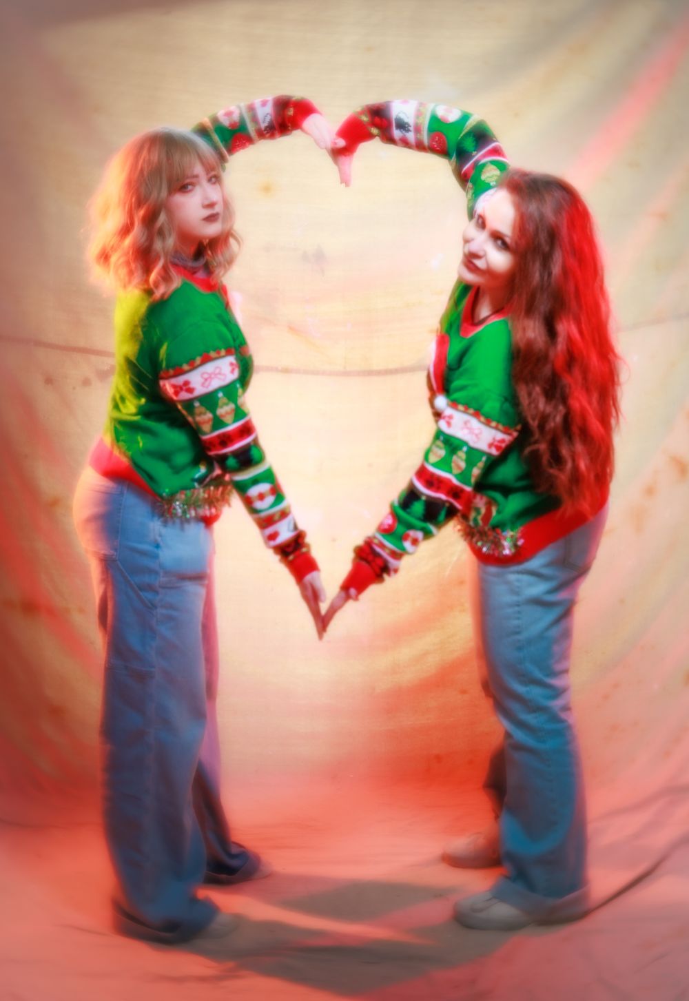 Two women in Christmas sweaters form a heart shape with their arms against a neutral backdrop.