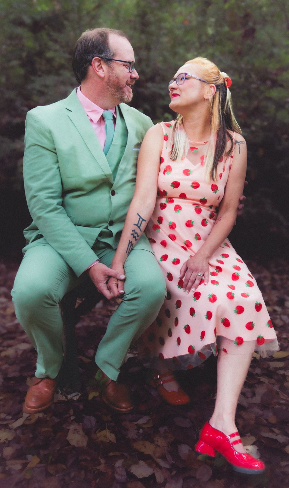 A couple sits outdoors in colorful retro outfits, man in green suit, woman in polka dot dress.