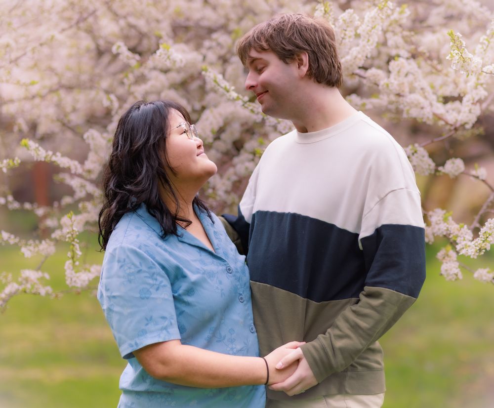 A couple holds hands and smiles at each other beneath blooming cherry blossom trees.