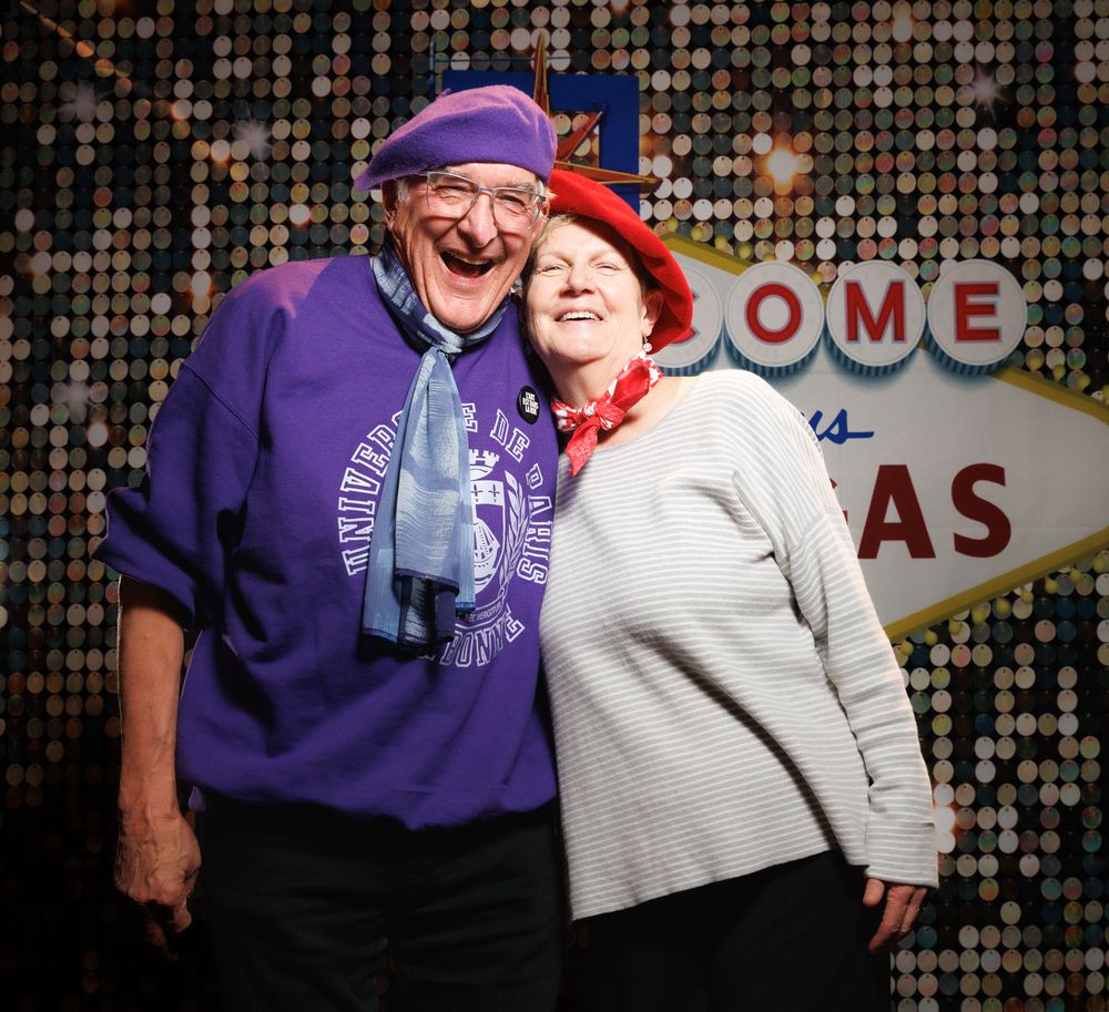 An elderly couple smiles and embraces in front of a Welcome to Las Vegas sign backdrop.