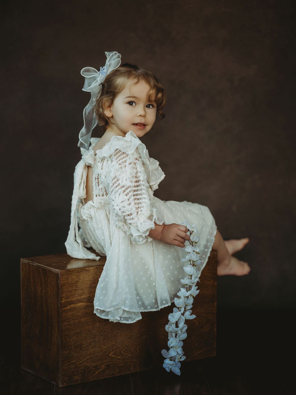 A child in a vintage white lace dress sits on a wooden box against a dark backdrop in a series of artistic portraits.