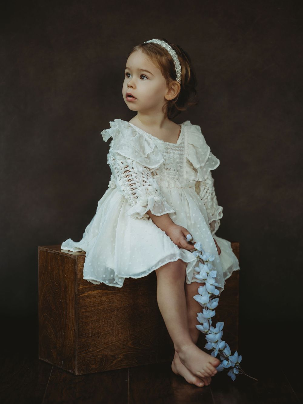 A small child in a vintage white lace dress sits on a wooden box against a dark backdrop with cascading flowers.