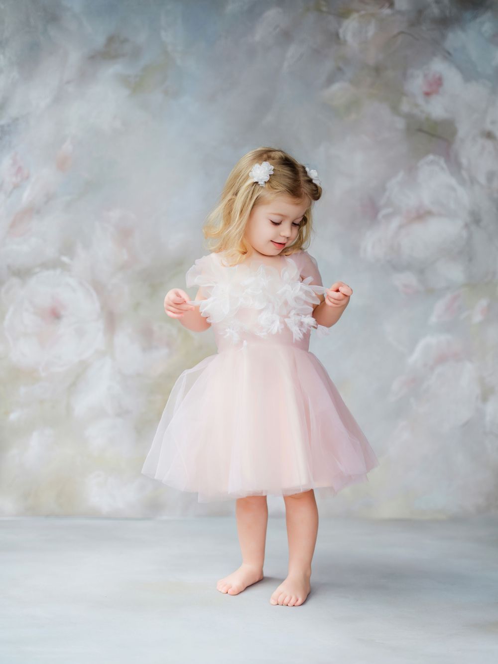 A toddler in a pink tulle dress twirls and plays in a dreamy studio setting with soft gray backdrop.