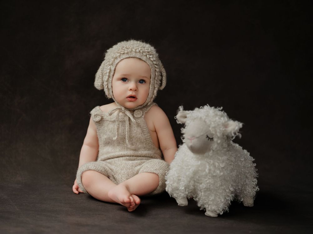 Baby in vintage-style cream dress and bonnet sits with white plush lamb toy against dark studio backdrop.