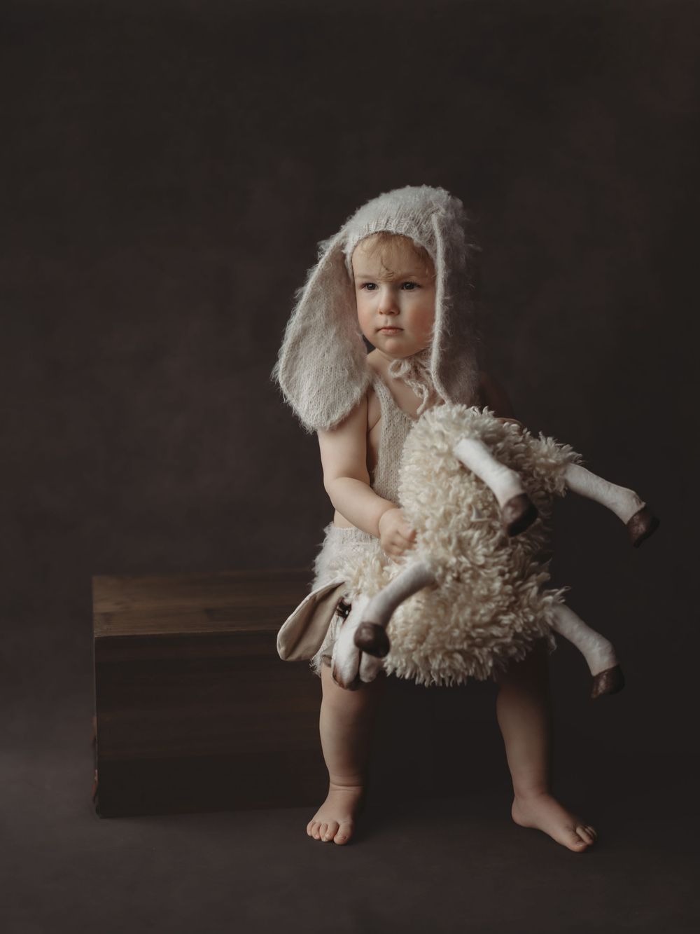 A small figure in white winter attire holds a plush sheep toy while standing barefoot against a dark background.