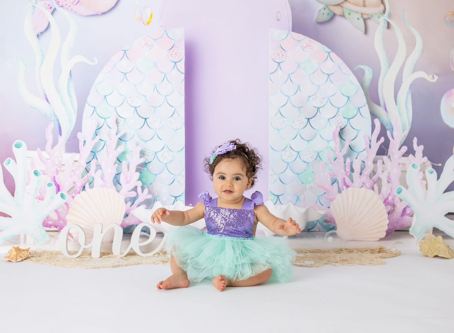 Baby in purple and mint tutu dress sitting against mermaid themed backdrop.