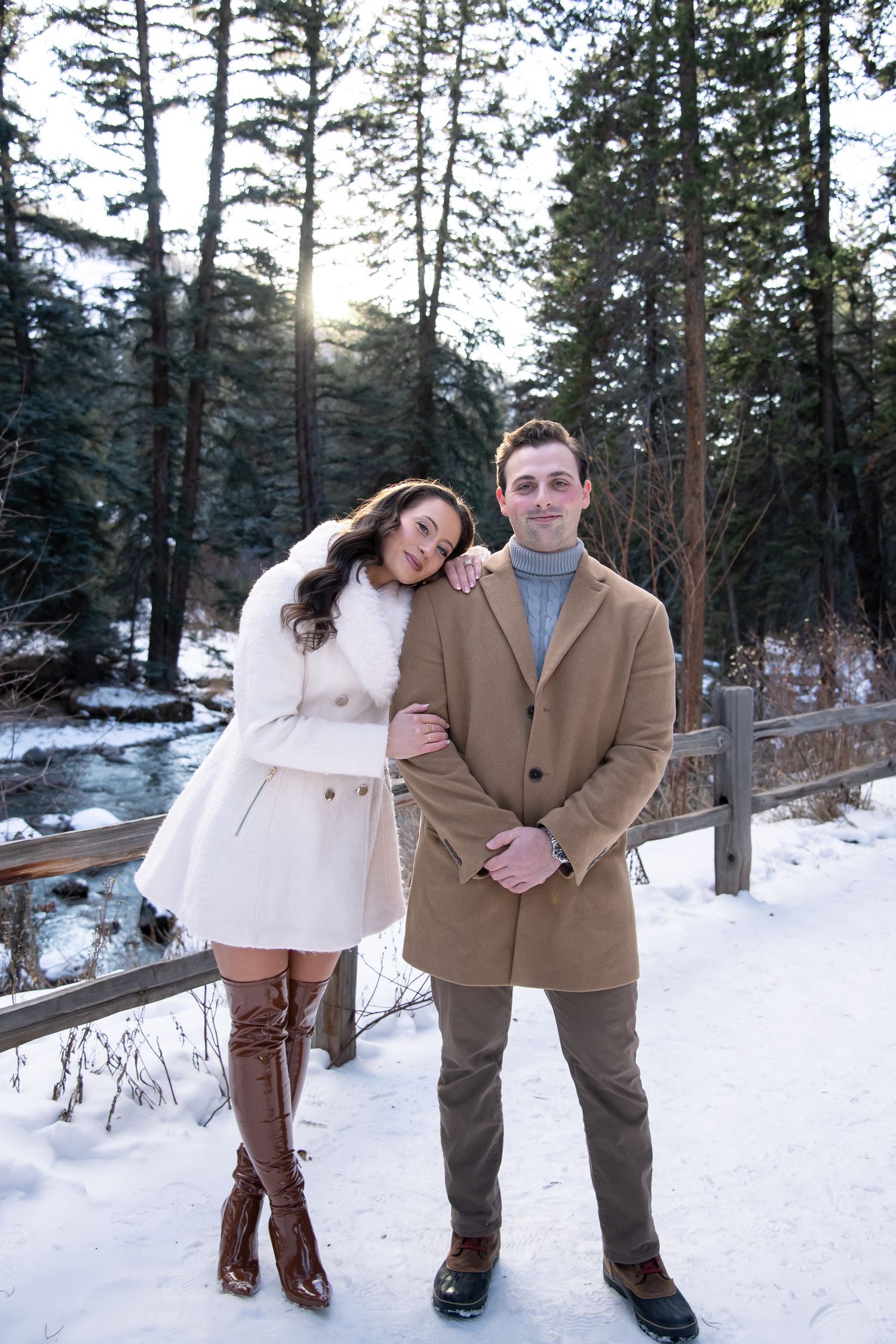 A cute couple poses together on a snowy winter path in the woods wearing stylish coats during their winter engagement session in Vail