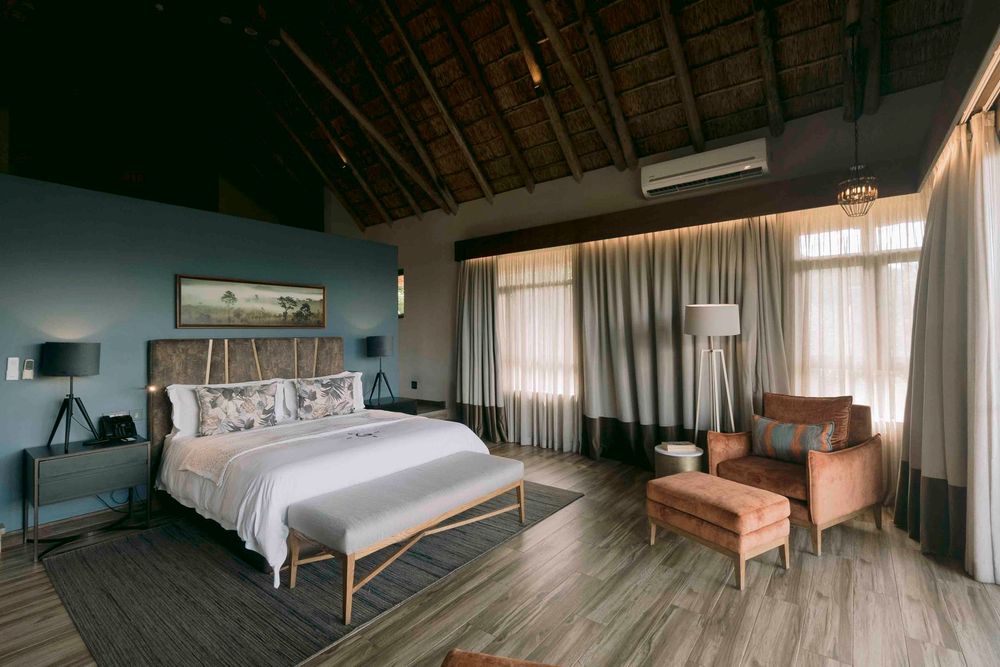 Spacious safari lodge bedroom with thatched ceiling, king bed, blue walls, and warm ambient lighting.