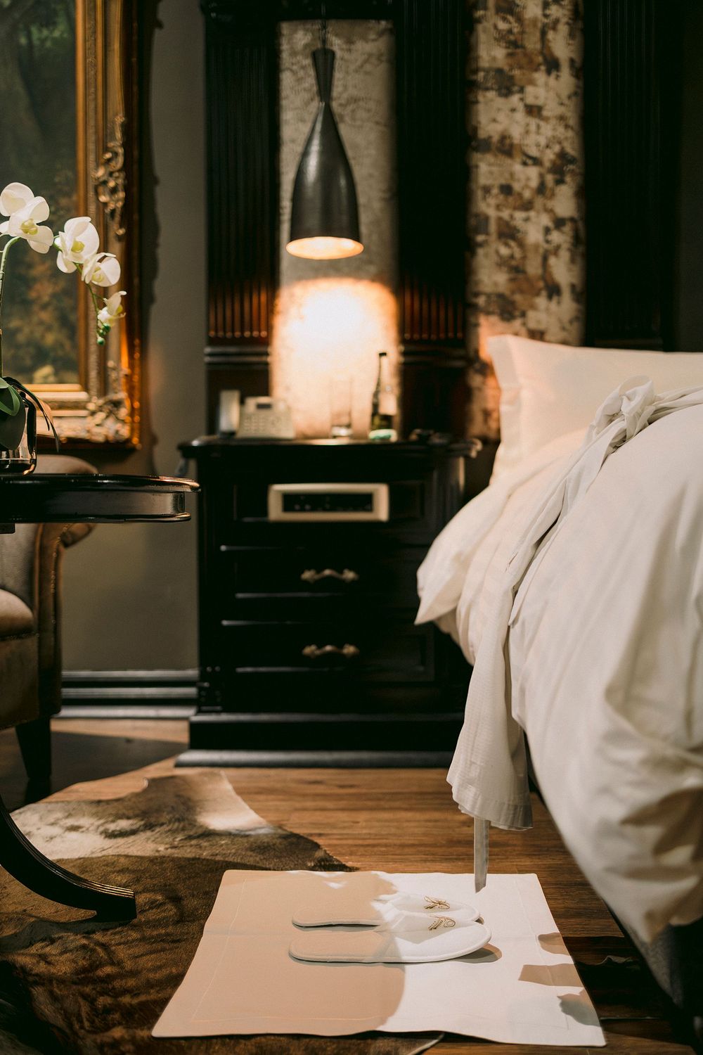 Elegant dark hotel room with white bedding, pendant lamp, orchids, and cowhide rug.