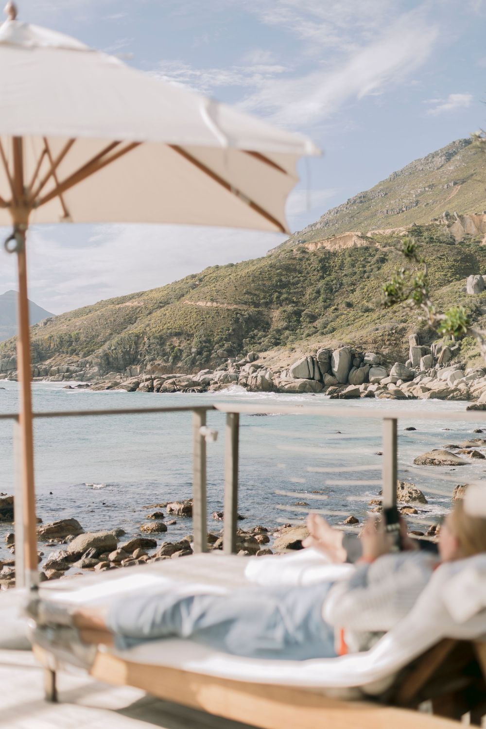 Person relaxing on a sun lounger under a white umbrella with a stunning rocky coastline view.