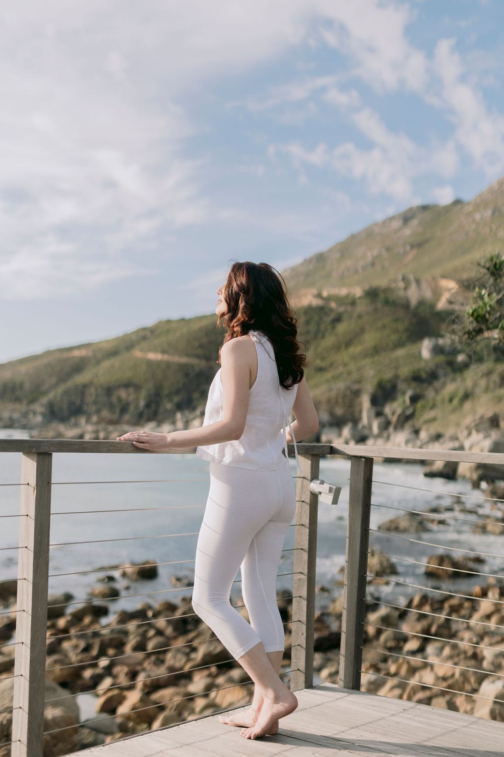 Barefoot woman in white outfit standing on a coastal deck overlooking rocky shores and green hills.