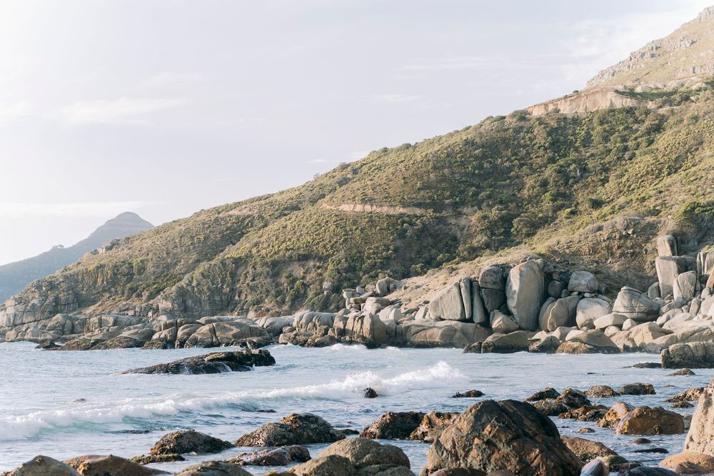 Dramatic rocky coastline with rolling waves and lush green hillside under a soft golden sky.