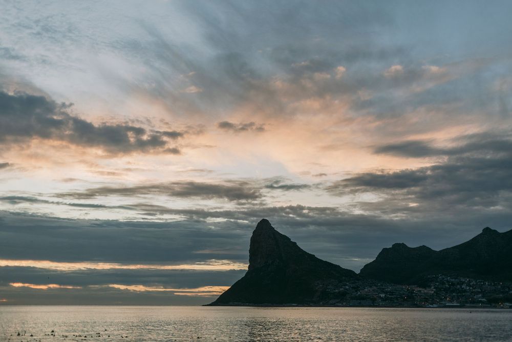 Moody dusk sky over tranquil bay with iconic mountain peak silhouette and distant town lights.