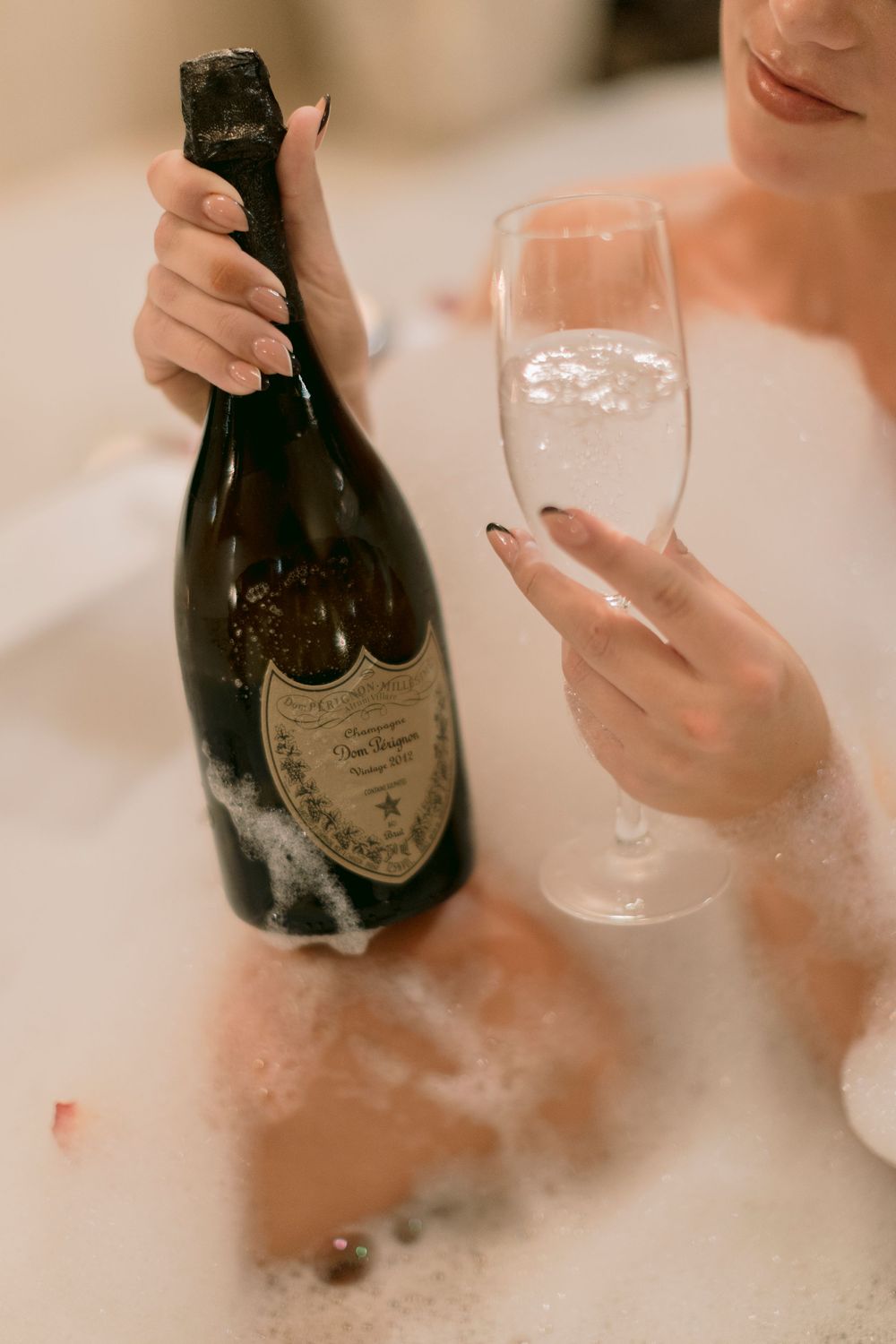 Woman relaxing in a bubble bath holding a bottle of Dom Perignon champagne and a crystal flute.