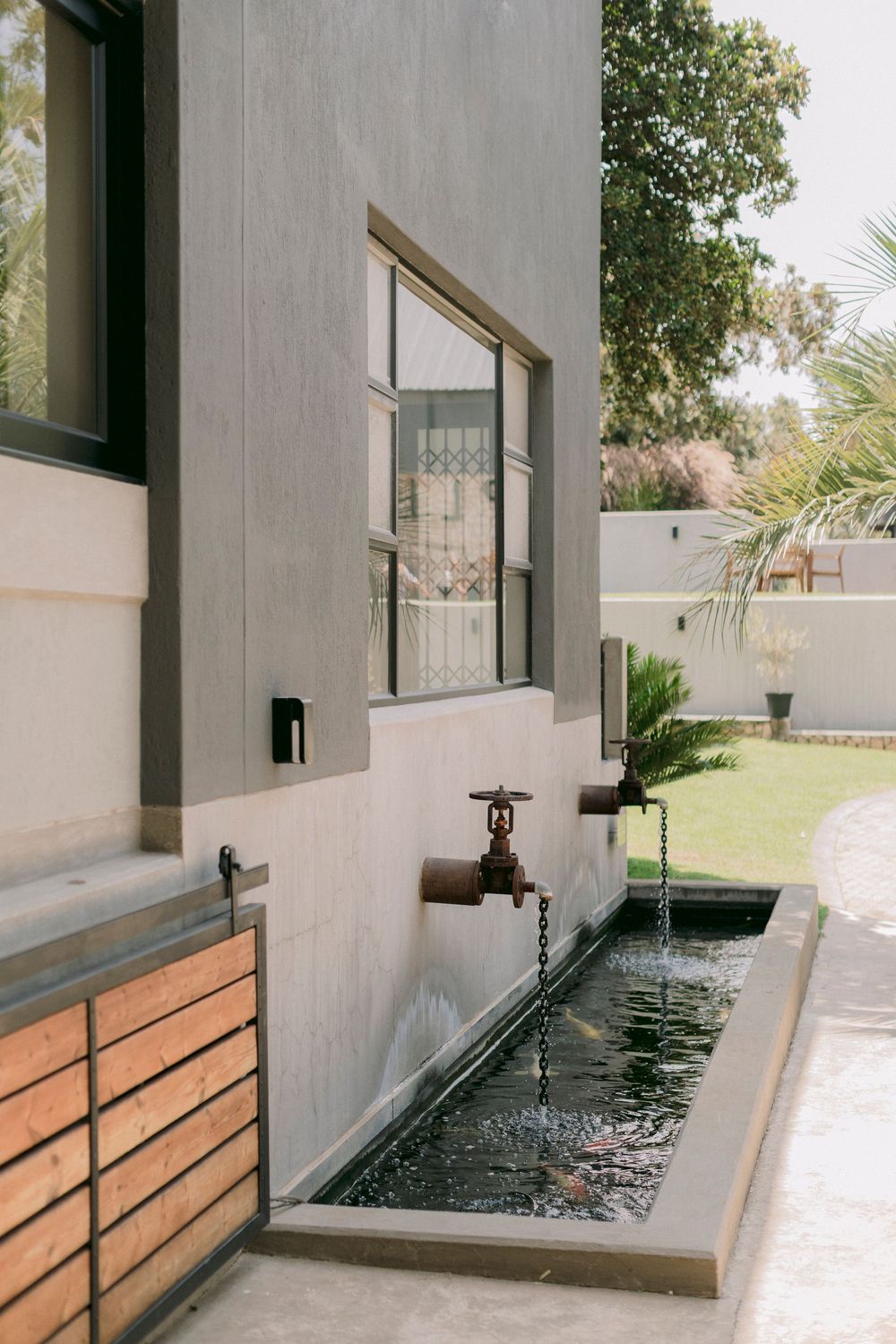 Rustic outdoor water feature with vintage faucets flowing into a narrow pool beside a modern grey stucco building.
