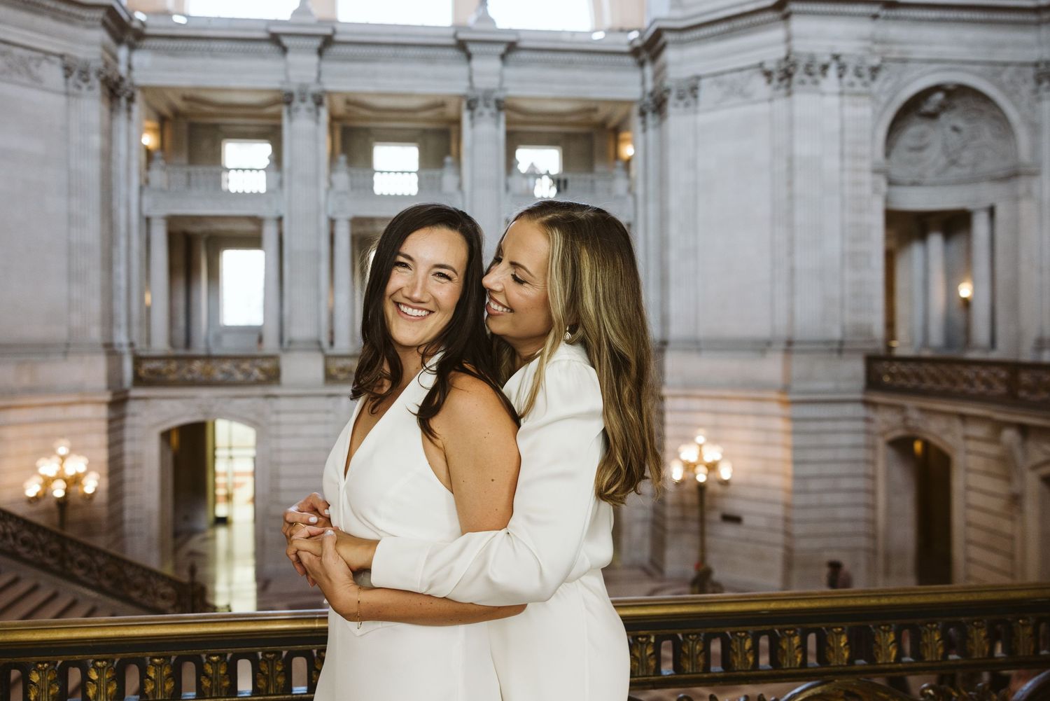 Two people in white dresses embrace and share joyful moments in a grand marble building interior.