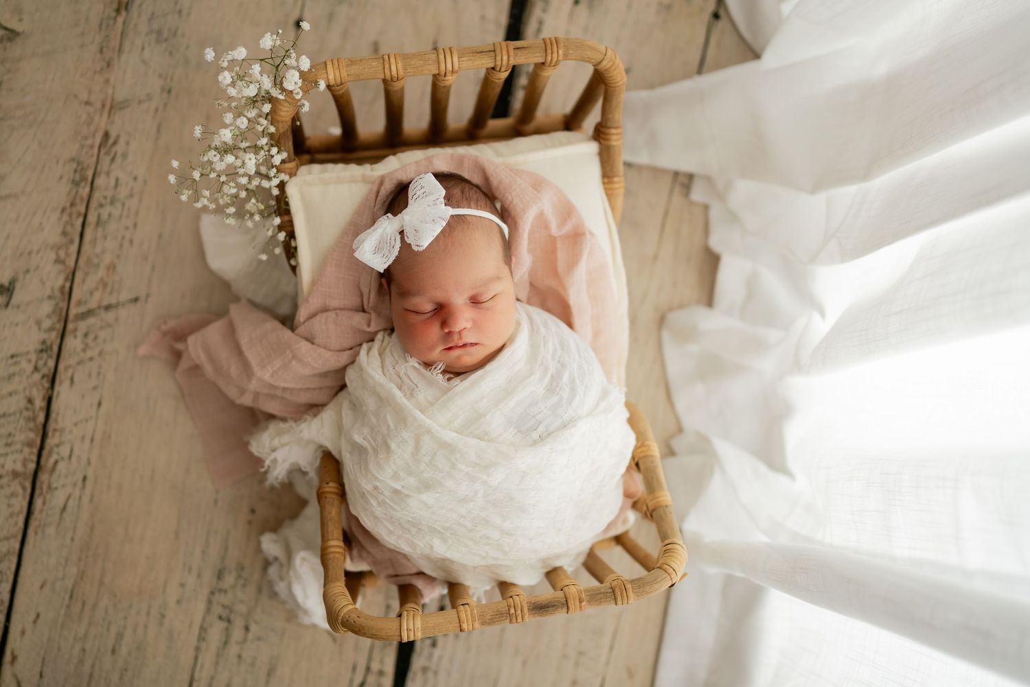 A newborn baby wearing white sleeps peacefully on a wooden basket lined with soft fabric in natural light.