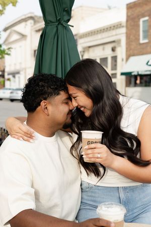 A couple shares an intimate moment while sitting outdoors at a cafe with green umbrellas visible in the background.