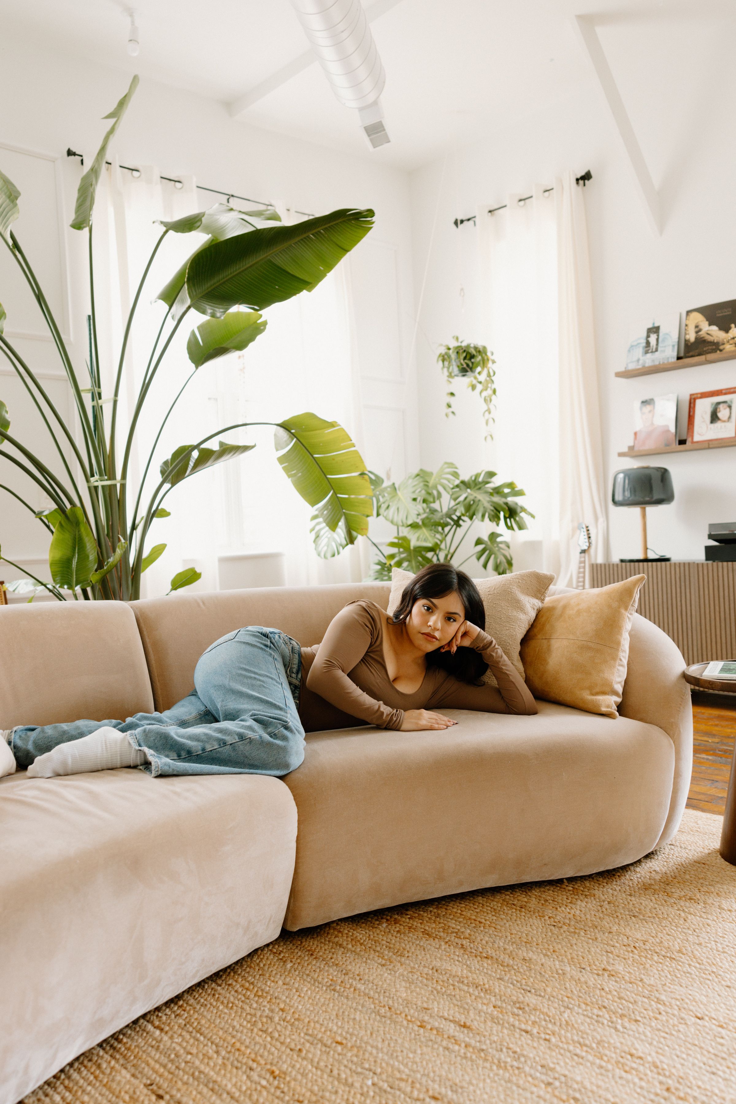 Cozy living room interior with large houseplants, beige sectional sofa, jute rug, and bright natural lighting.