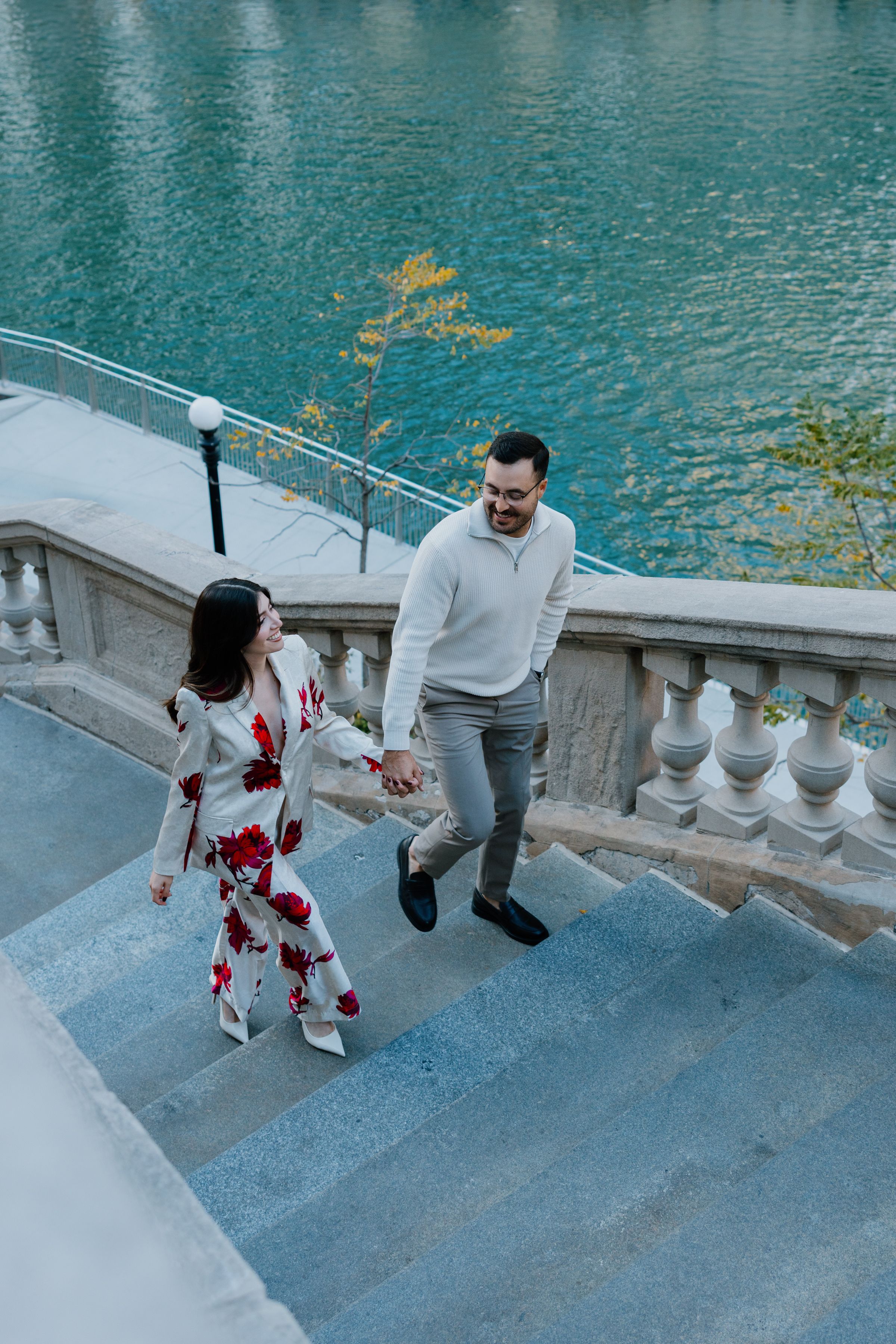 A couple walks up stone steps by turquoise water, with one person wearing a floral dress and another in casual grey attire.