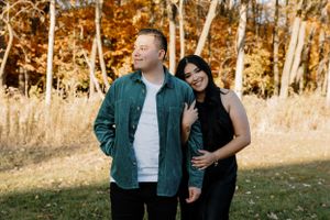 A couple stands together in an autumn setting with golden trees and grass in the background.