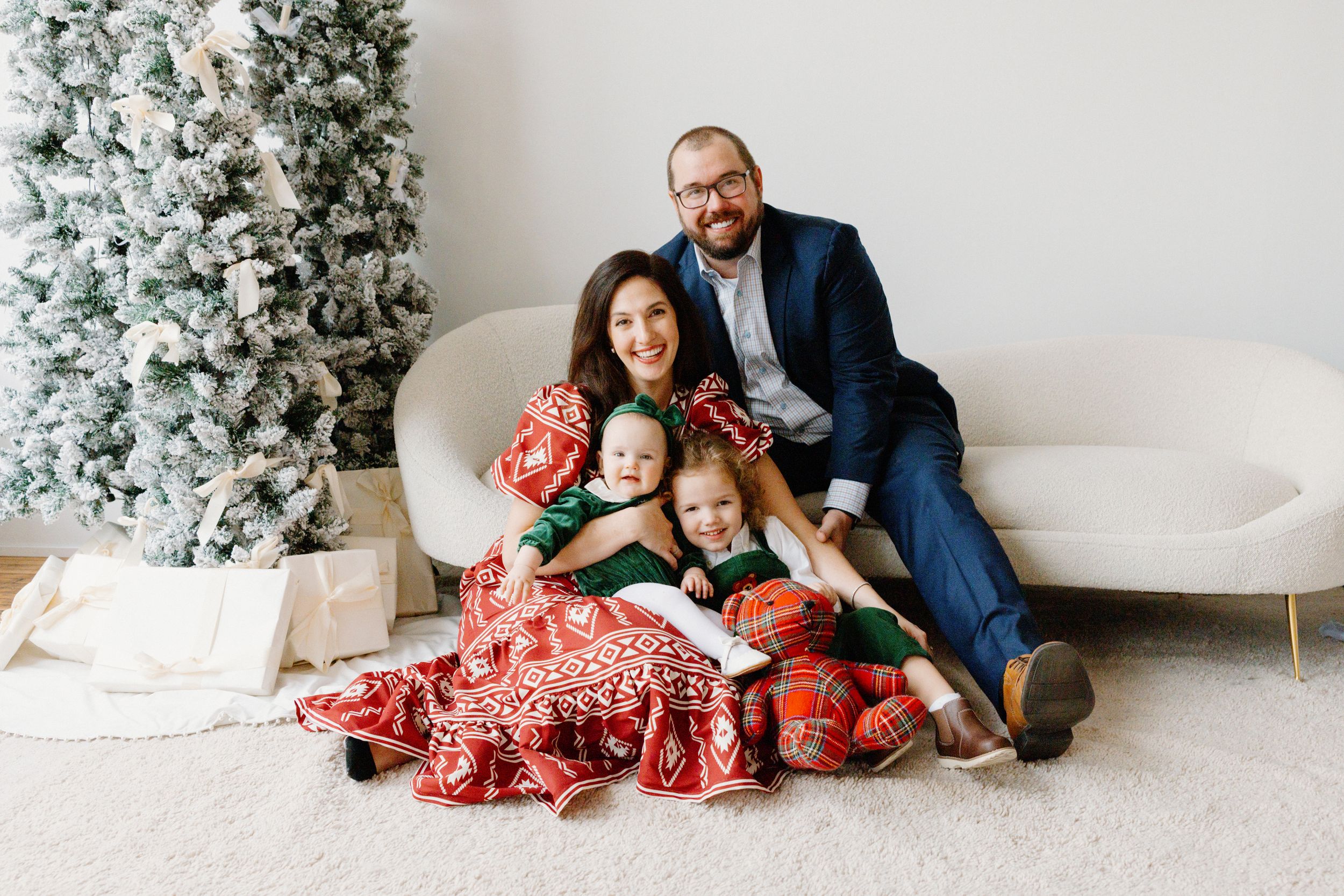A family sits together on the floor near a decorated white Christmas tree, wearing coordinated red holiday outfits.
