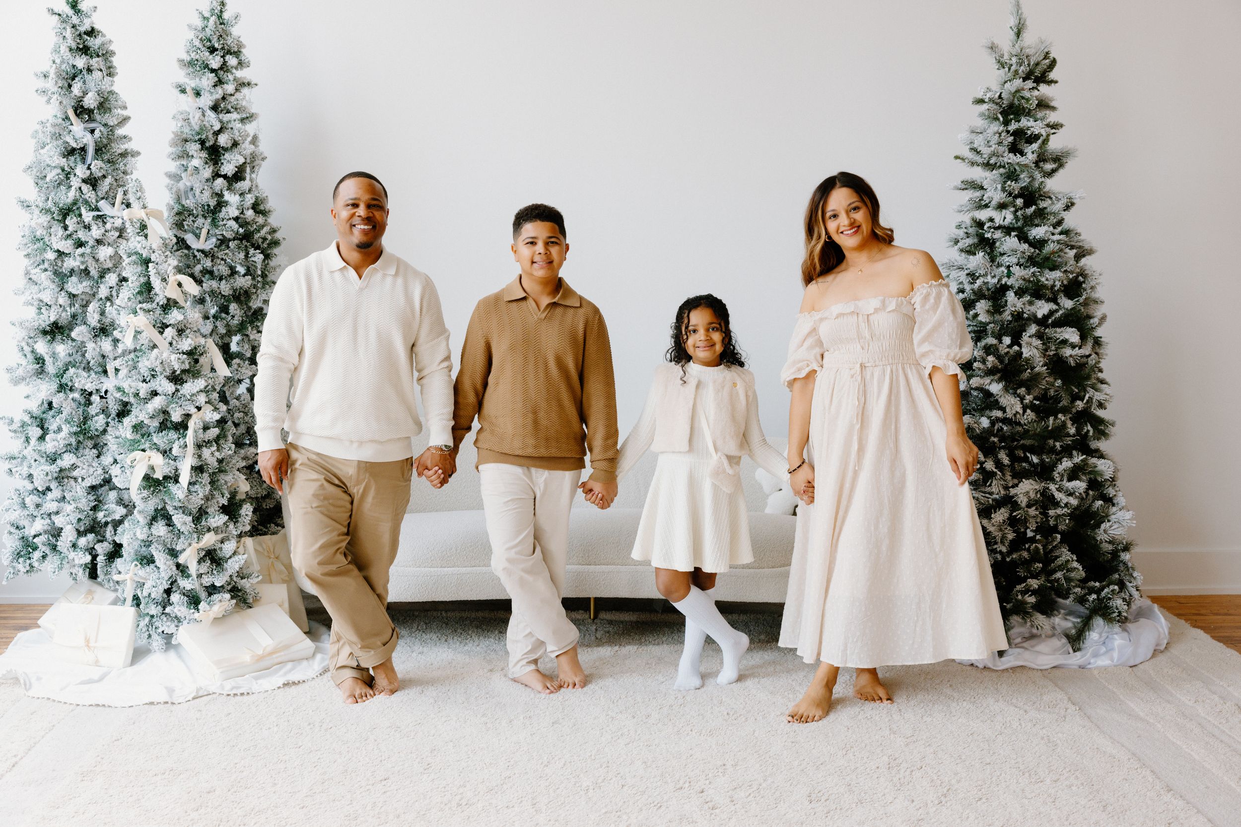 People dressed in coordinating beige and white outfits pose together in front of snow-covered Christmas trees in a studio.