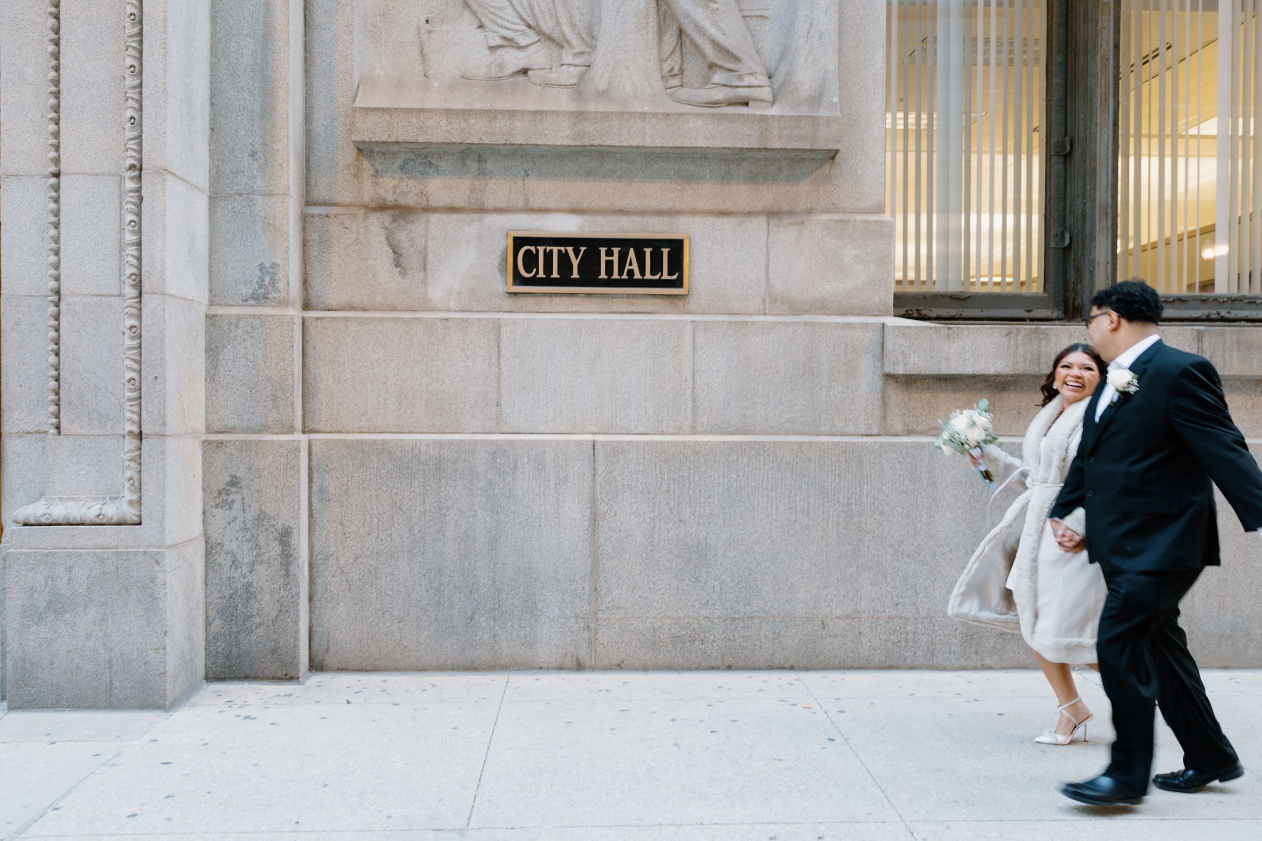 Two figures in formal attire stand together outside a city hall building with classical stone architecture.