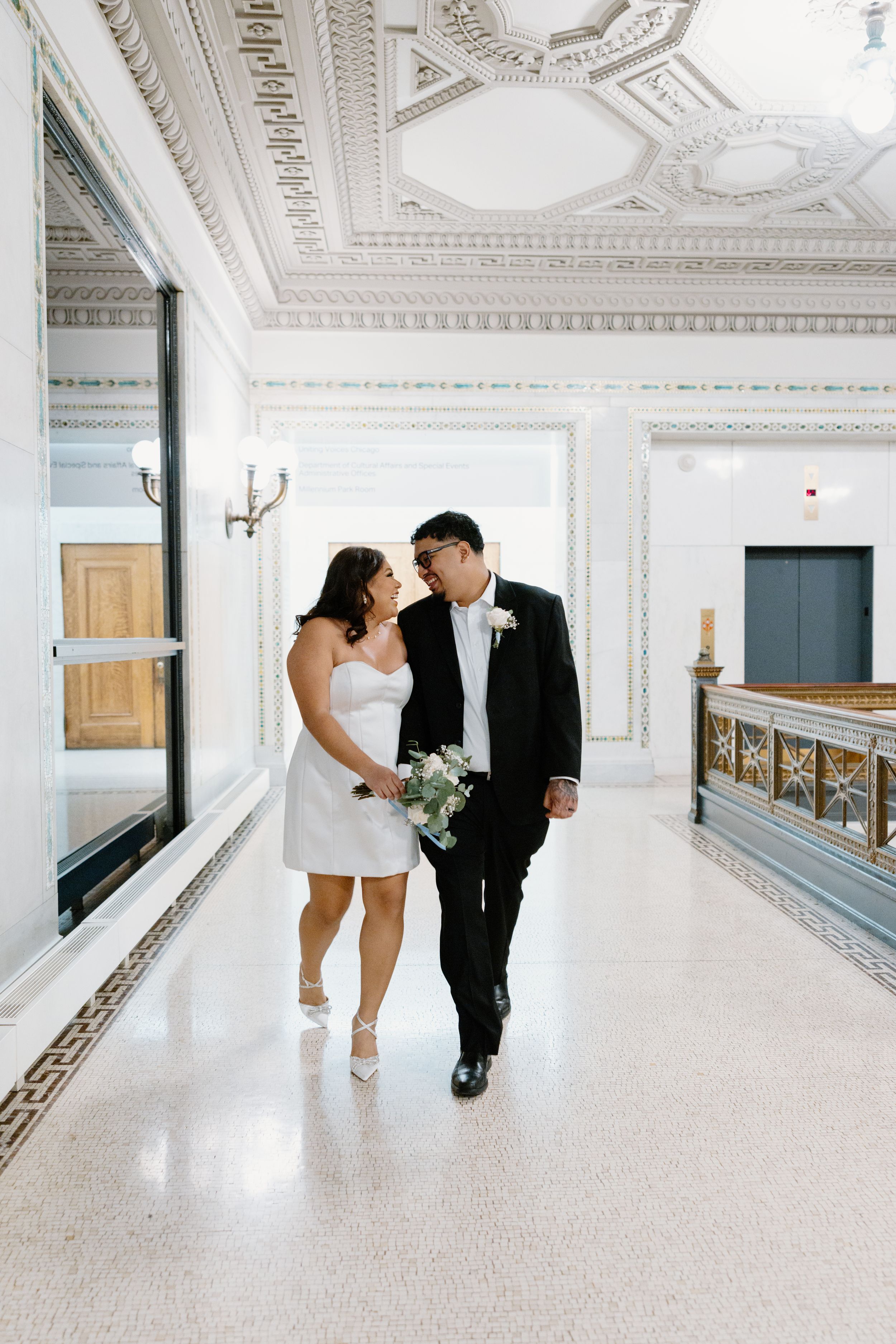 A couple walks down an elegant hallway with ornate white moldings and a decorative ceiling in a historic building.