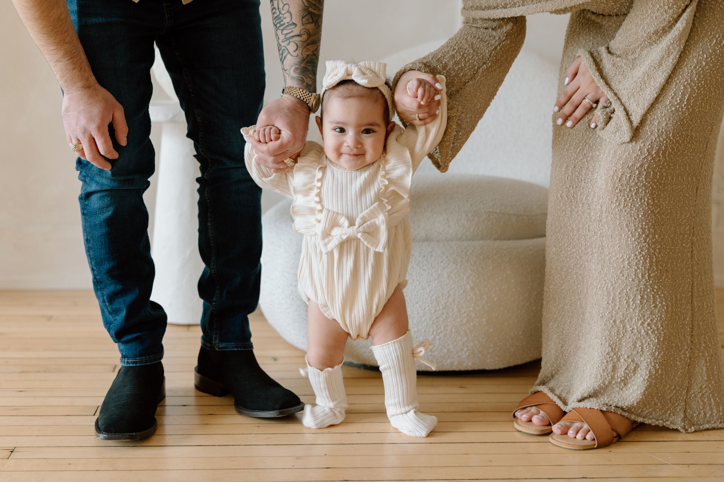 A baby taking first steps in a white romper while being supported by parents on hardwood floors.