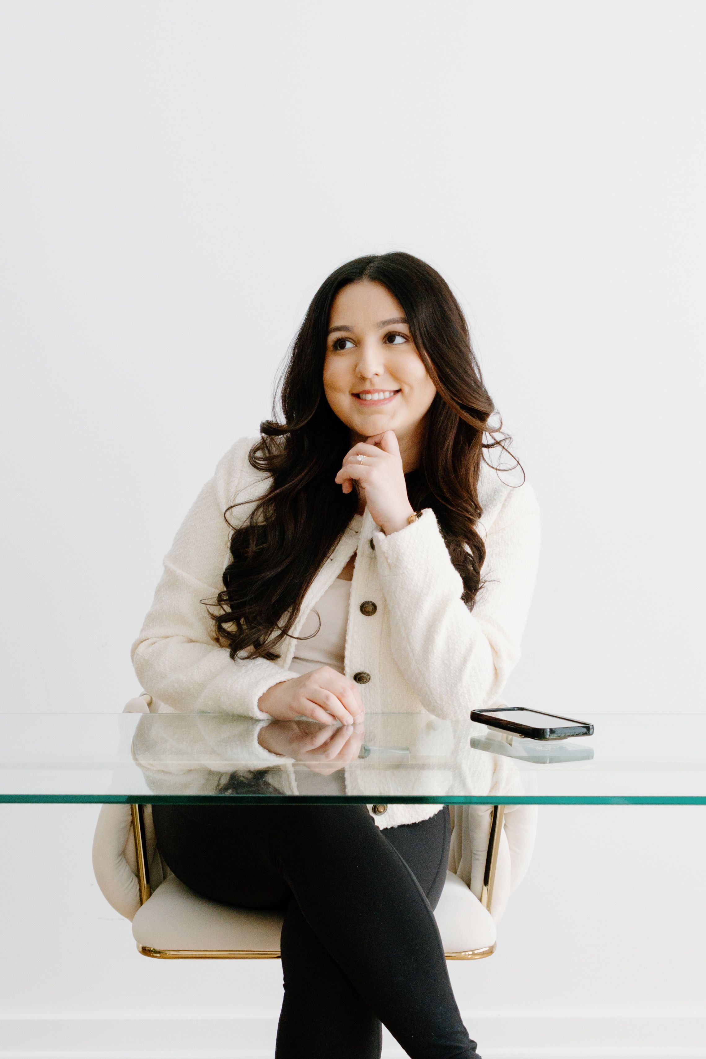 A person in a cream-colored button-up cardigan sits thoughtfully at a glass desk with a modern minimalist aesthetic.