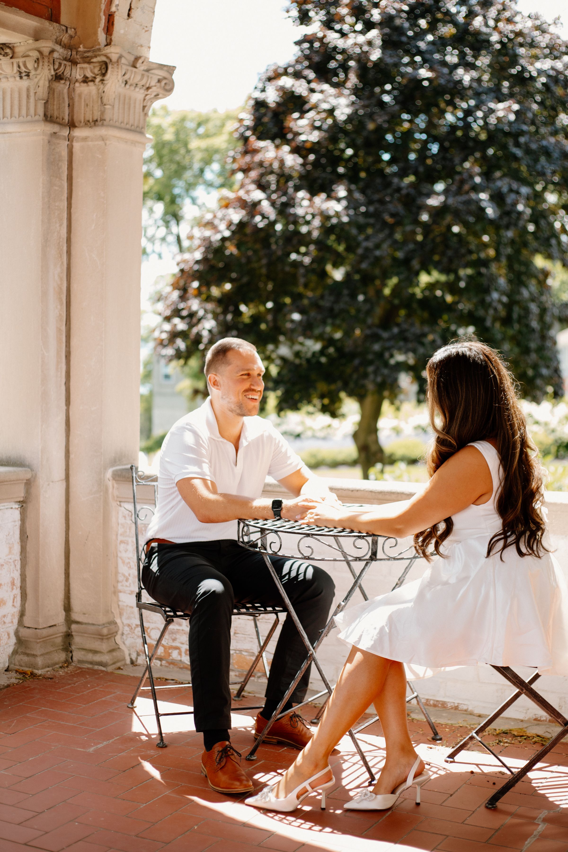 Couple having coffee at outdoor patio cafe with ornate pillars and trees in background on sunny day.