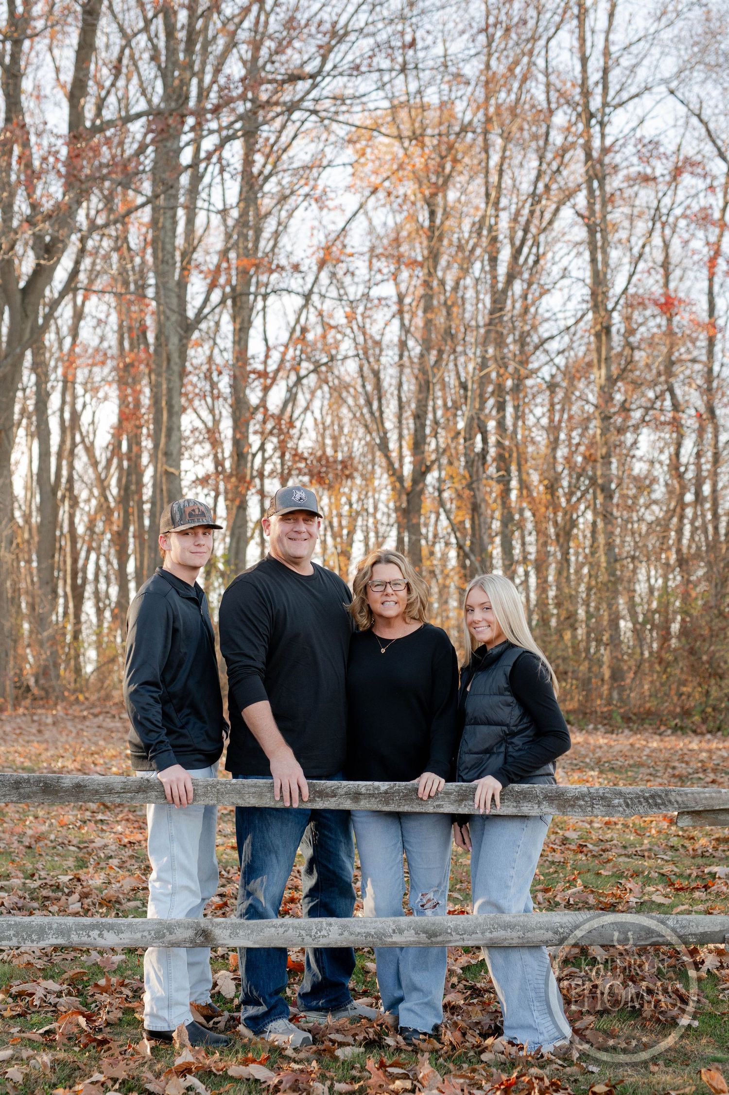 Family group stands together at rustic wooden fence during fall with bare trees behind them.