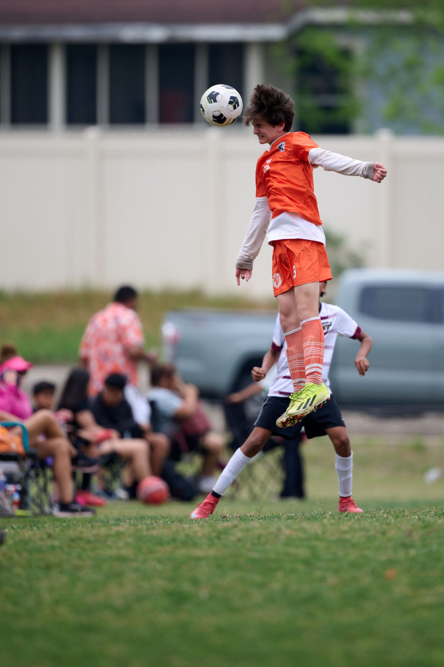 A soccer player in orange jersey jumps high to head the ball during a match with spectators watching.