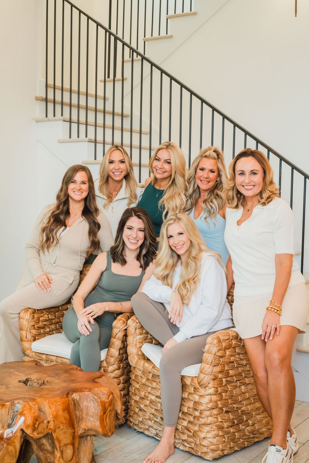 A group of women in casual attire posing together on woven ottomans by a staircase in a bright interior setting.