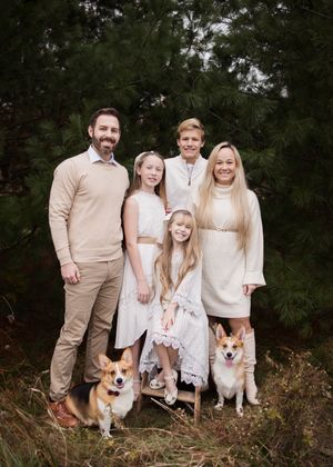 A family wearing coordinated beige and white outfits poses outdoors with their Corgi dog in a natural setting.