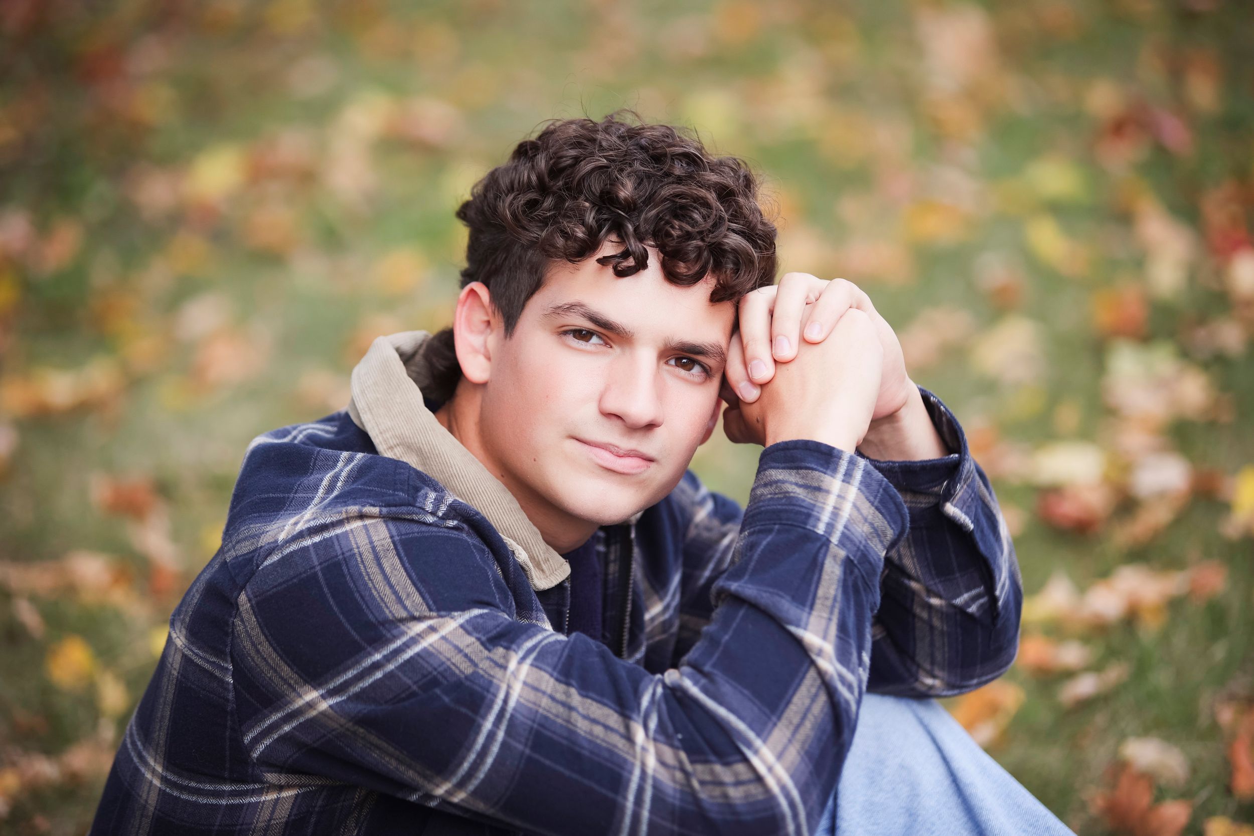A person in a navy plaid shirt poses outdoors with autumn leaves scattered on the ground.