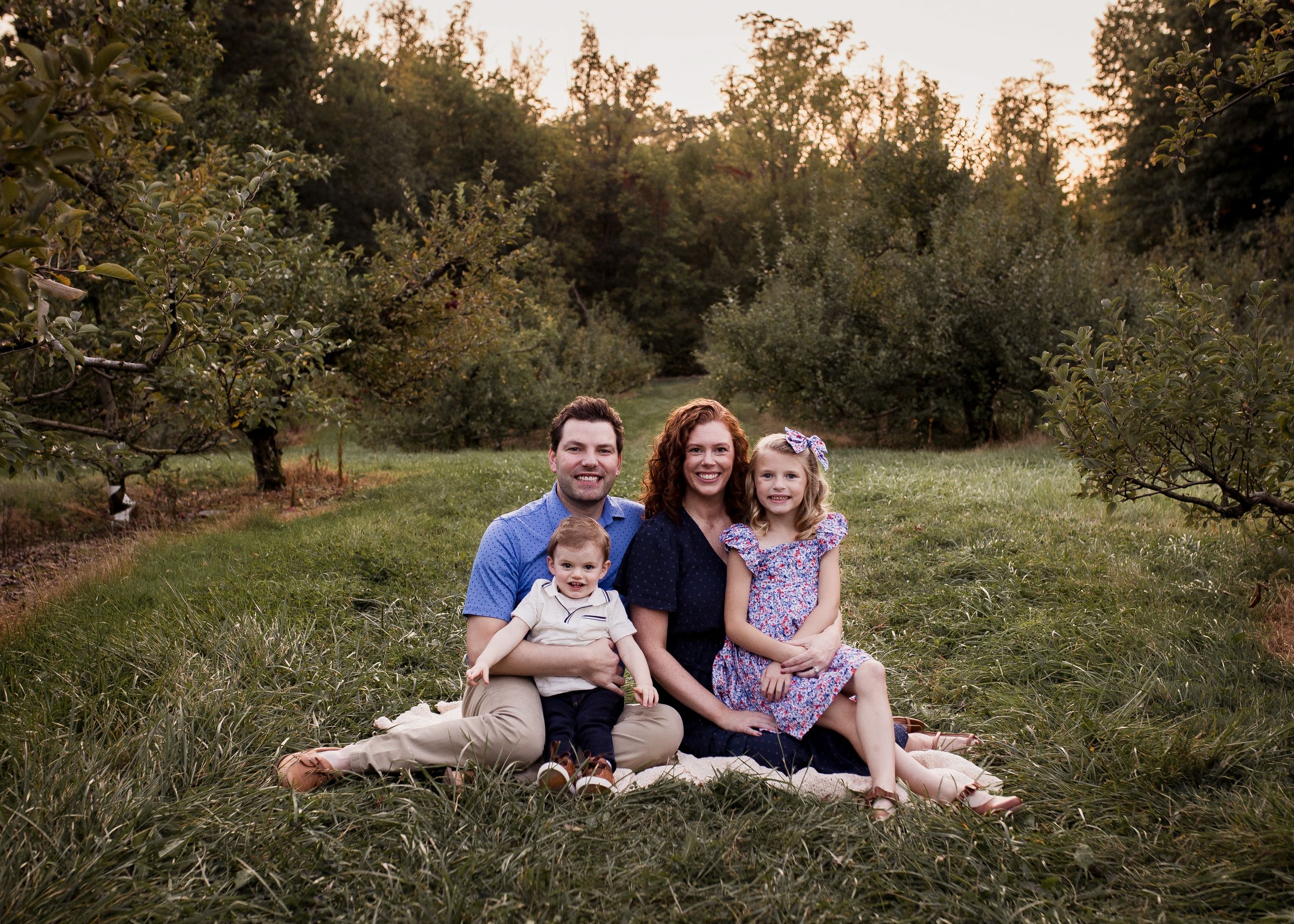A family sits together on a blanket in an orchard during golden hour at sunset.