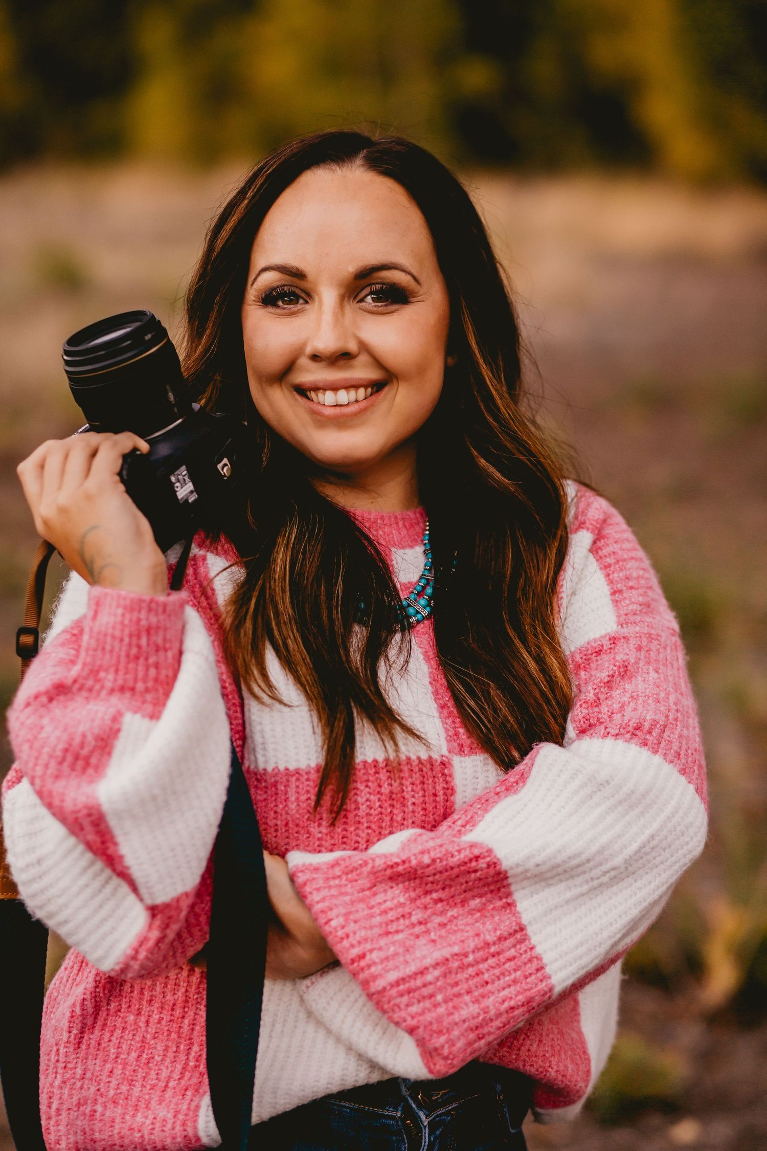 Someone wearing a pink and white striped sweater holds a camera while smiling outdoors.