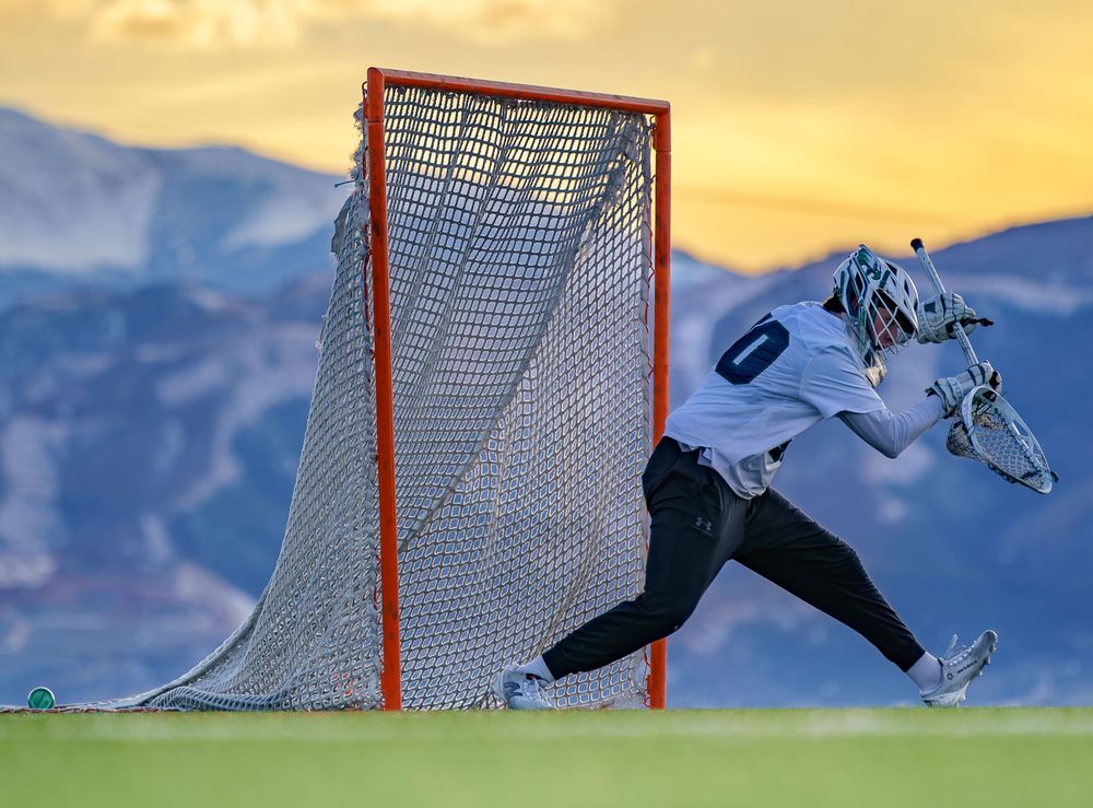 Lacrosse goalie makes dramatic save against sunset sky with mountain backdrop.