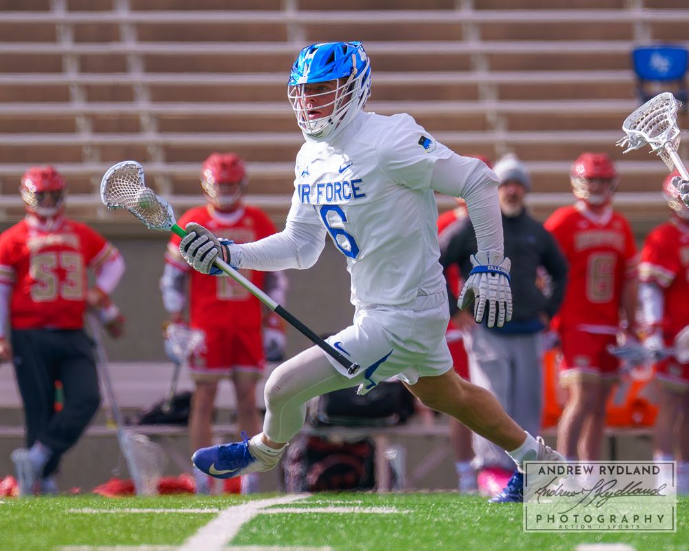 Lacrosse player in light blue uniform carries ball while running across field during game.