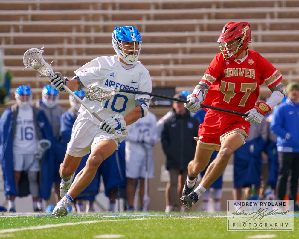 Lacrosse players from opposing teams sprint down field during competitive match.