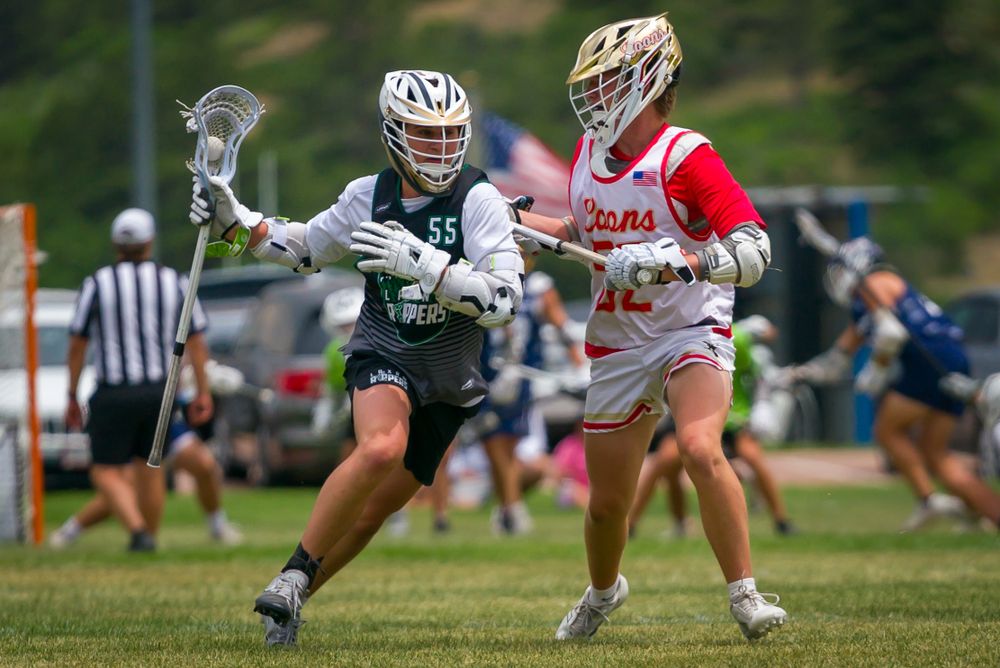 Two lacrosse players in green and red uniforms compete for ball control during match.