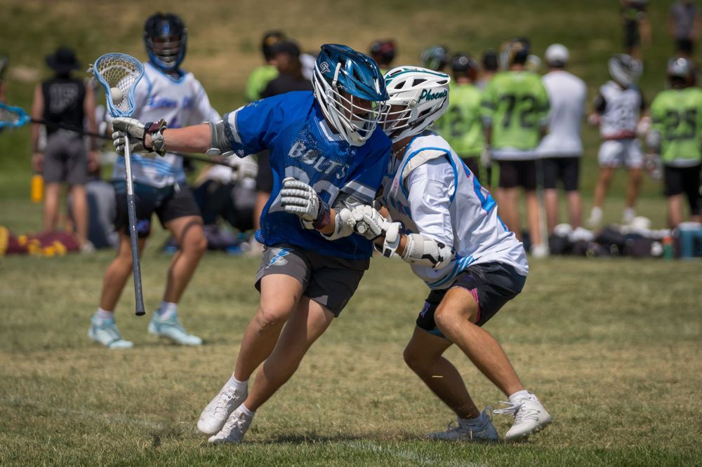Two lacrosse players battle for ball possession during outdoor match on grass field.