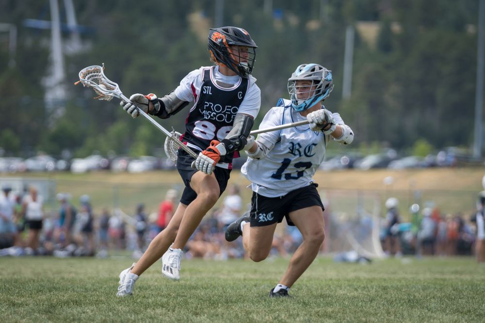 Two youth lacrosse players compete for ball possession during outdoor tournament match.