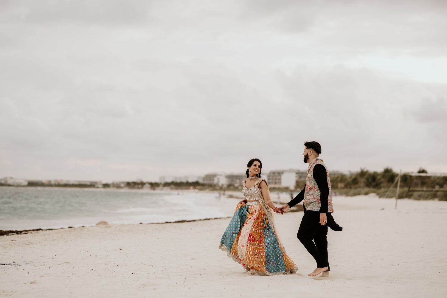 A couple in formal attire walks together on a sandy beach during an overcast day by the ocean.