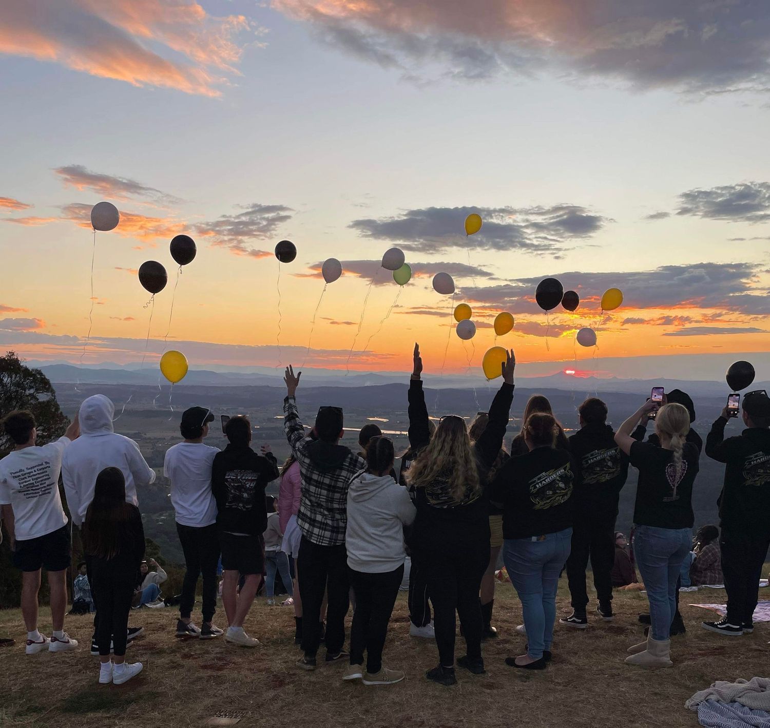 Crowd releasing balloons into colorful sunset sky during outdoor gathering.
